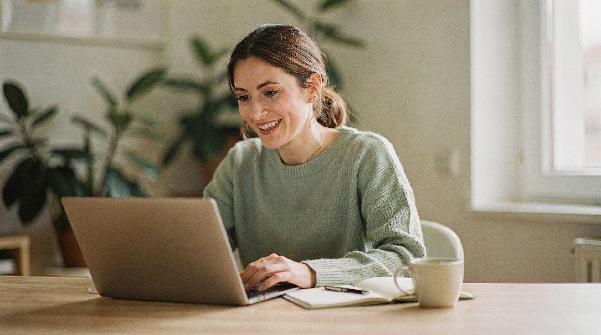 A doctor in a soft knit jumper smiling on a video call at her laptop in a bright home study.