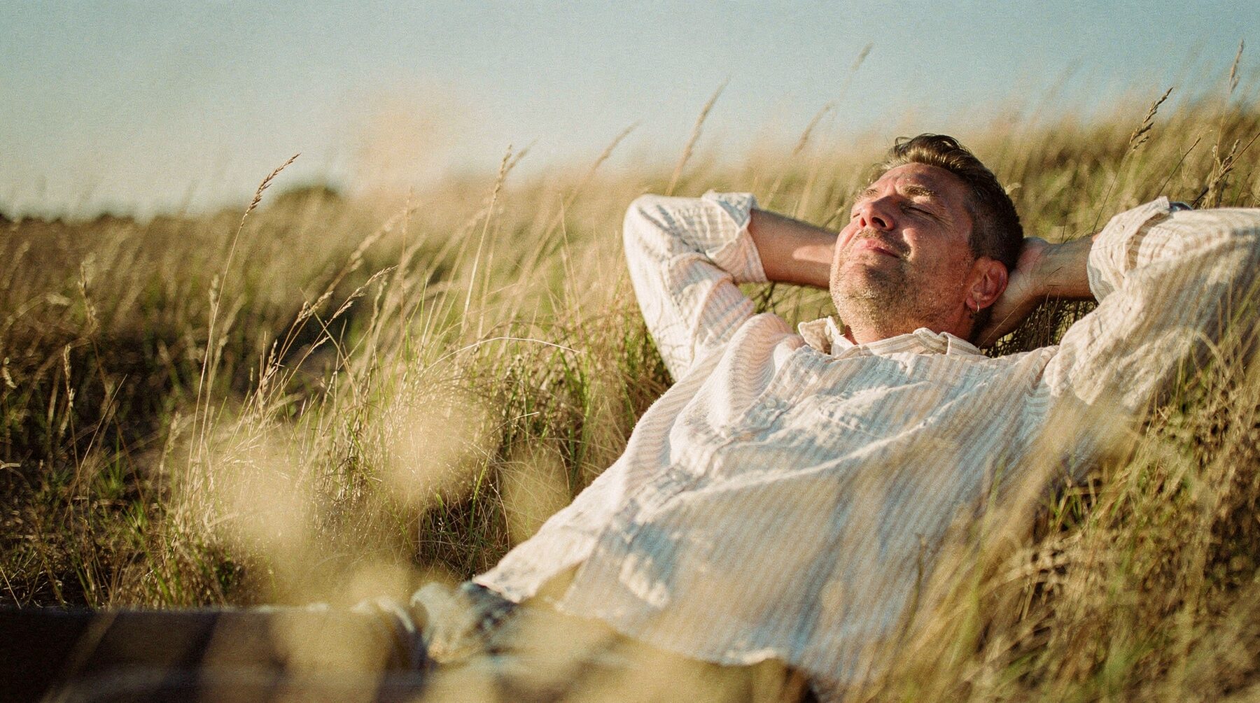 A man lies back in long summer grass, hands behind his head, eyes closed in the sun.