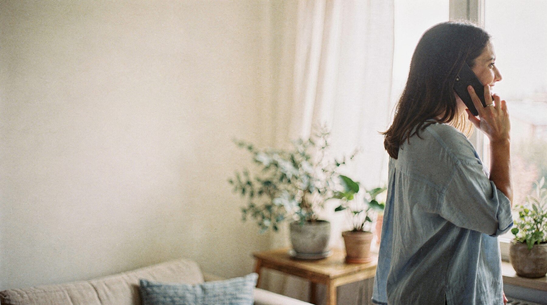A woman at home by a bright window, talking on the phone and smiling.